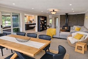 A modern living room with a fireplace, a dining area with a wooden table, and a kitchen in the background. - Sunny Coastal Escape with Pool and Stunning Ocean Views! (Whangarei)