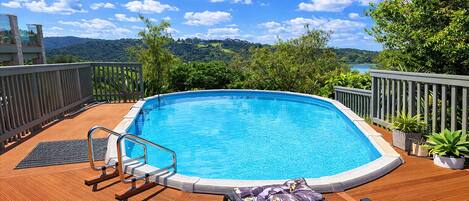 A swimming pool with a wooden deck, lounge chairs, and a view of a lake and hills.