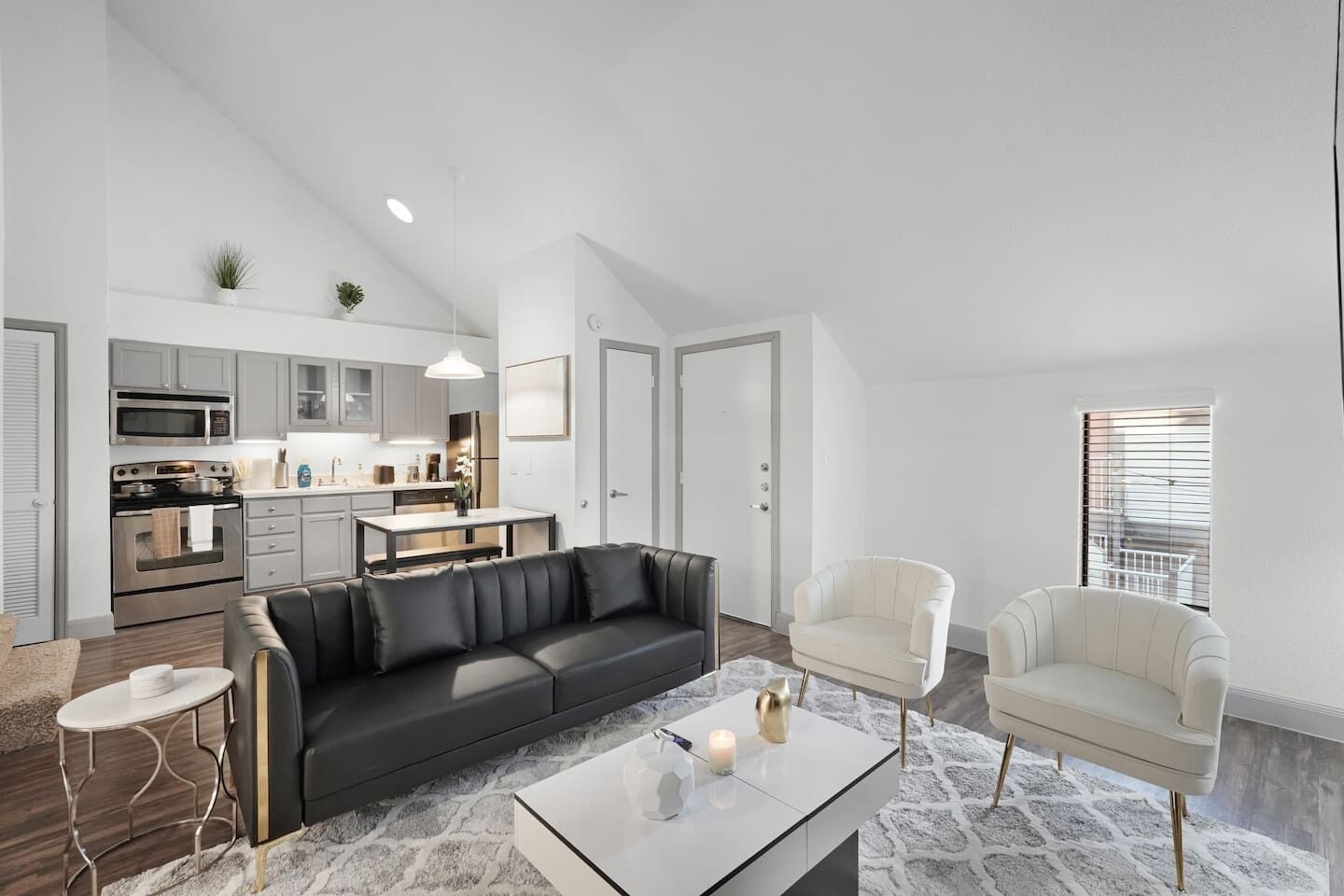 A modern living space with a black leather sofa, a white armchair, a glass coffee table, and a kitchen area with grey cabinets and stainless steel appliances.