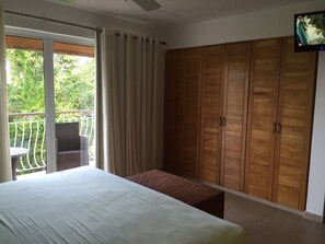 A bedroom with a bed, a wooden wardrobe, a balcony with a table and chairs, and a television on the wall.