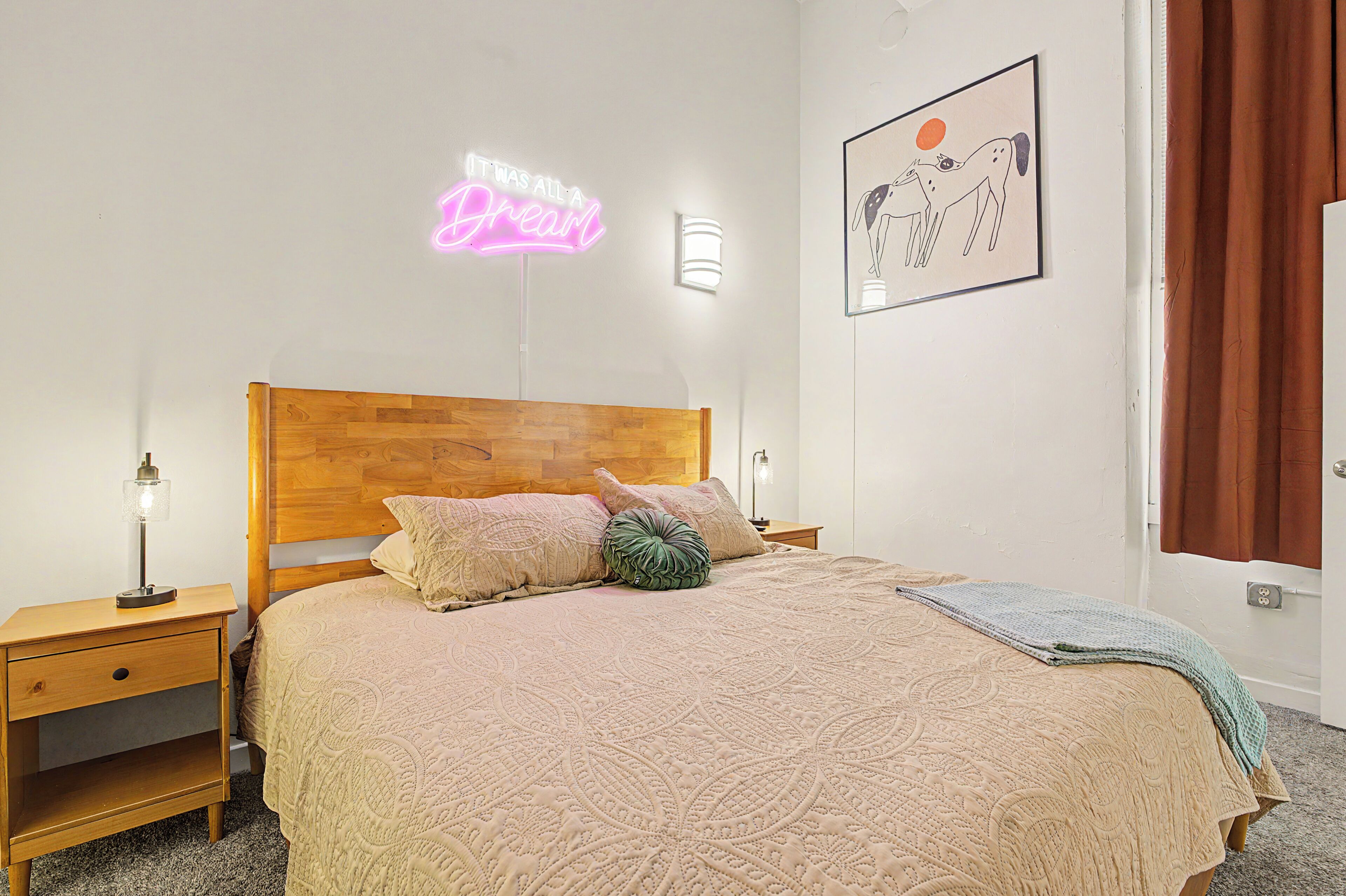 A neatly arranged bedroom with a wooden headboard, a patterned bedspread, and a neon sign on the wall.