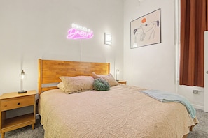 Un dormitorio ordenado con cabecera de madera, colcha estampada y un letrero de neón en la pared.