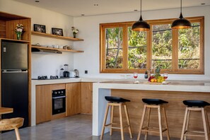 A modern kitchen with wooden cabinetry, a built-in oven, a refrigerator, and a large window with a view of greenery. - Villa LIMA 2 Uluwatu (Pecatu)