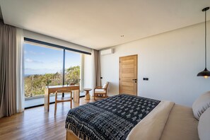 A bedroom with a large bed, a desk, and a chair, a wooden door, and a view through a large window. - Villa LIMA 2 Uluwatu (Pecatu)