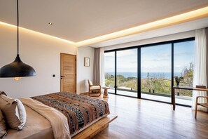 A bedroom with a large bed, a wooden door, a chair, a small table, and a view of the outdoors. - Villa LIMA 2 Uluwatu (Pecatu)