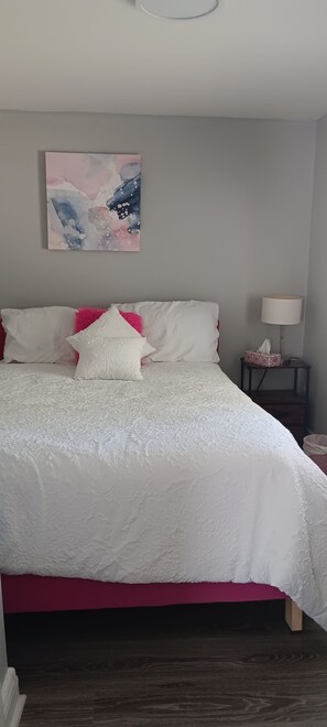 A bedroom with a bed, pillows, a nightstand, a lamp, and a painting on the wall. - Your Windsor Home Away from Home | 3BR One-Storey (Windsor)