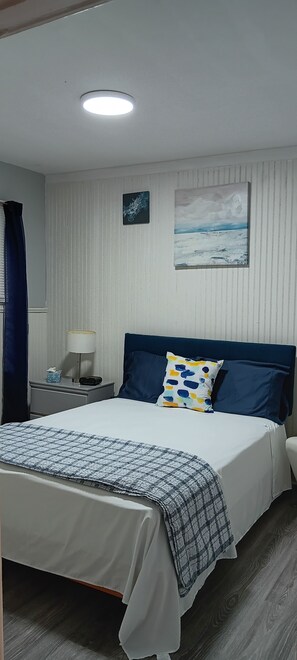 A hotel room with a bed, a nightstand, a lamp, a painting, and a curtain. - Your Windsor Home Away from Home | 3BR One-Storey (Windsor)