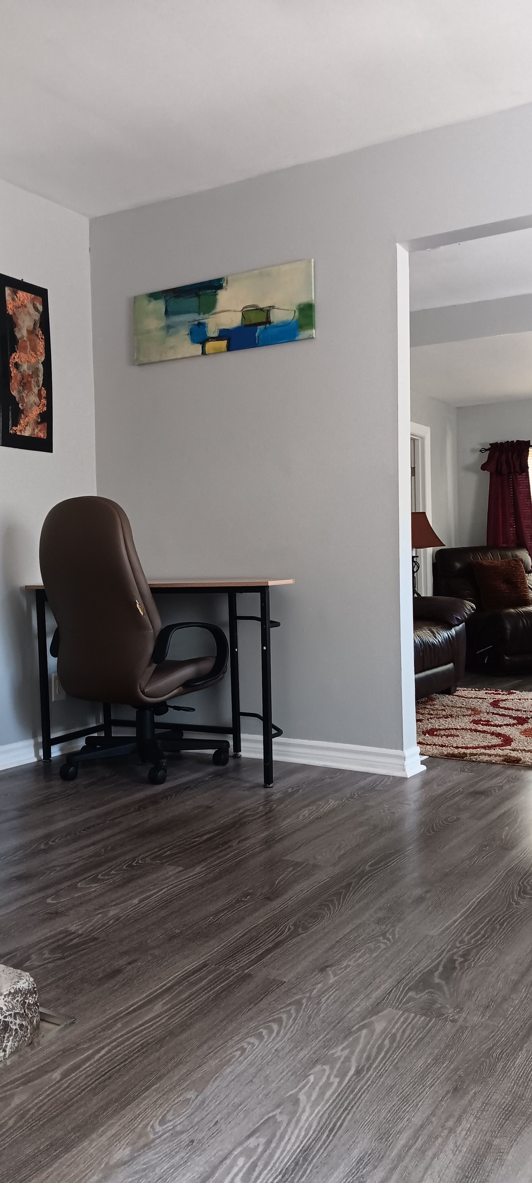 Ein Zimmer mit Schreibtisch, Stuhl, einem Bild an der Wand und Blick in einen weiteren Raum.