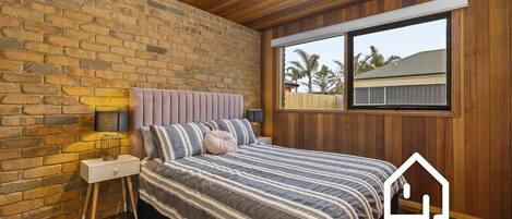 A bedroom with a brick wall, wooden ceiling, a bed with striped bedding, a bedside table with a lamp, and a window with a view of palm trees.