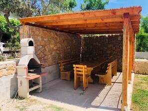 Een terras buiten met een stenen muur, een houten tafel en banken, en een bakoven.