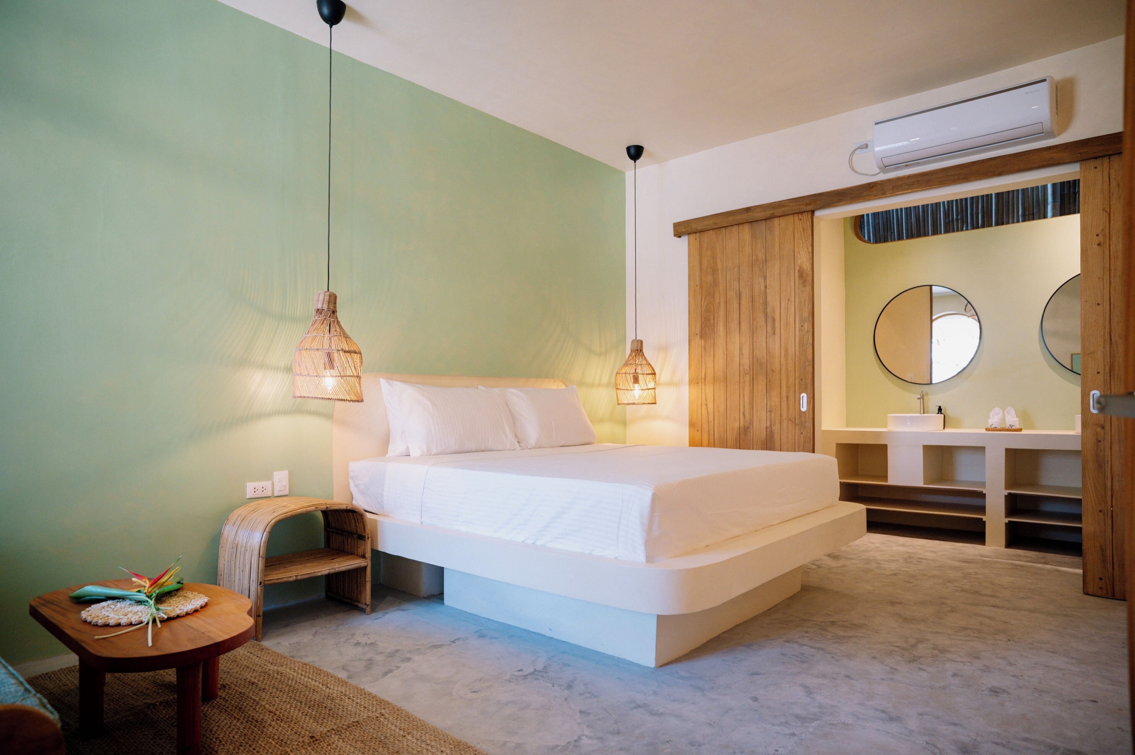 A bedroom with a bed, bedside lamps, a wooden wardrobe, a bathroom with a sink and mirror, and an air conditioning unit.