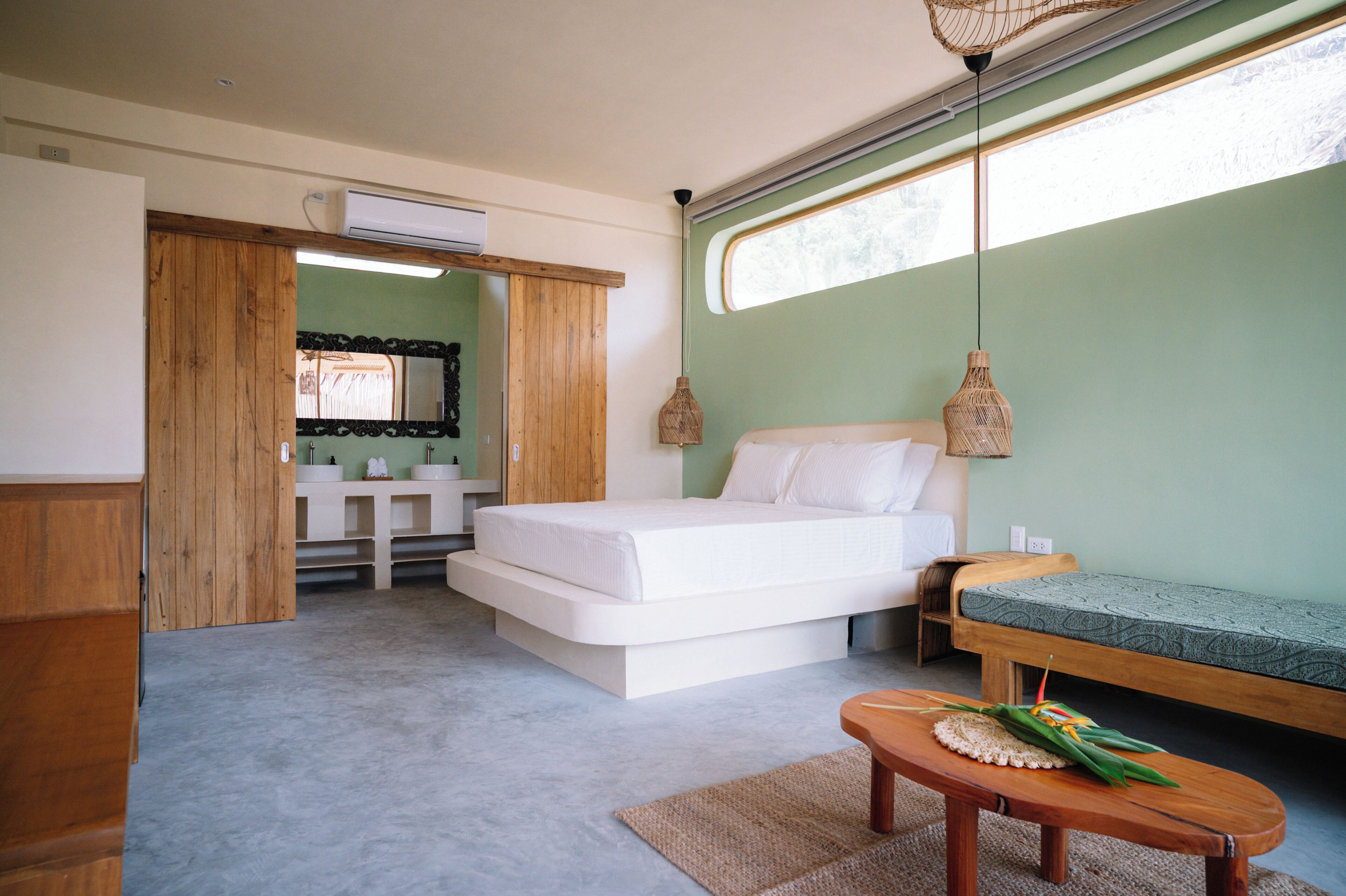 A bedroom with a bed, a wooden bench, a small table, a window with a view, and a bathroom with a mirror and sink.