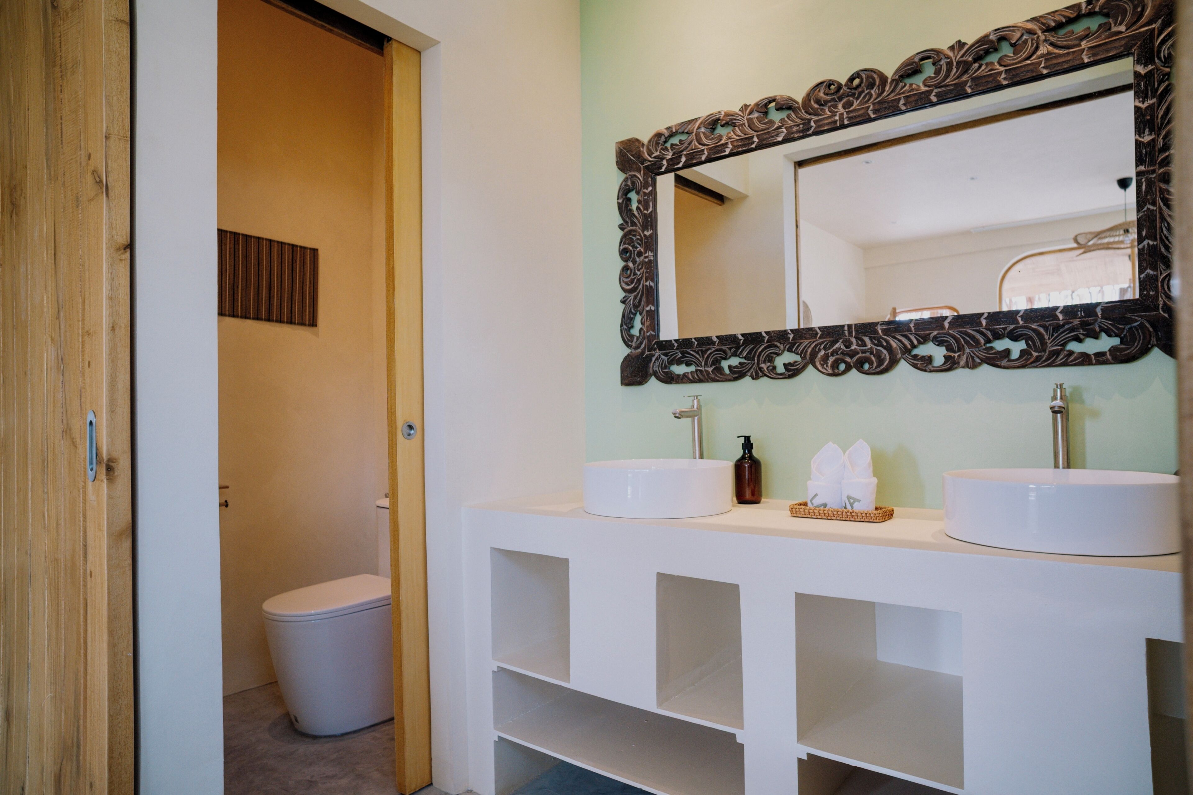 A bathroom with a wooden door, a white toilet, and a vanity with two sinks and a mirror.