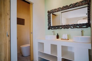 Un baño con puerta de madera, inodoro blanco y tocador con dos lavamanos y un espejo.