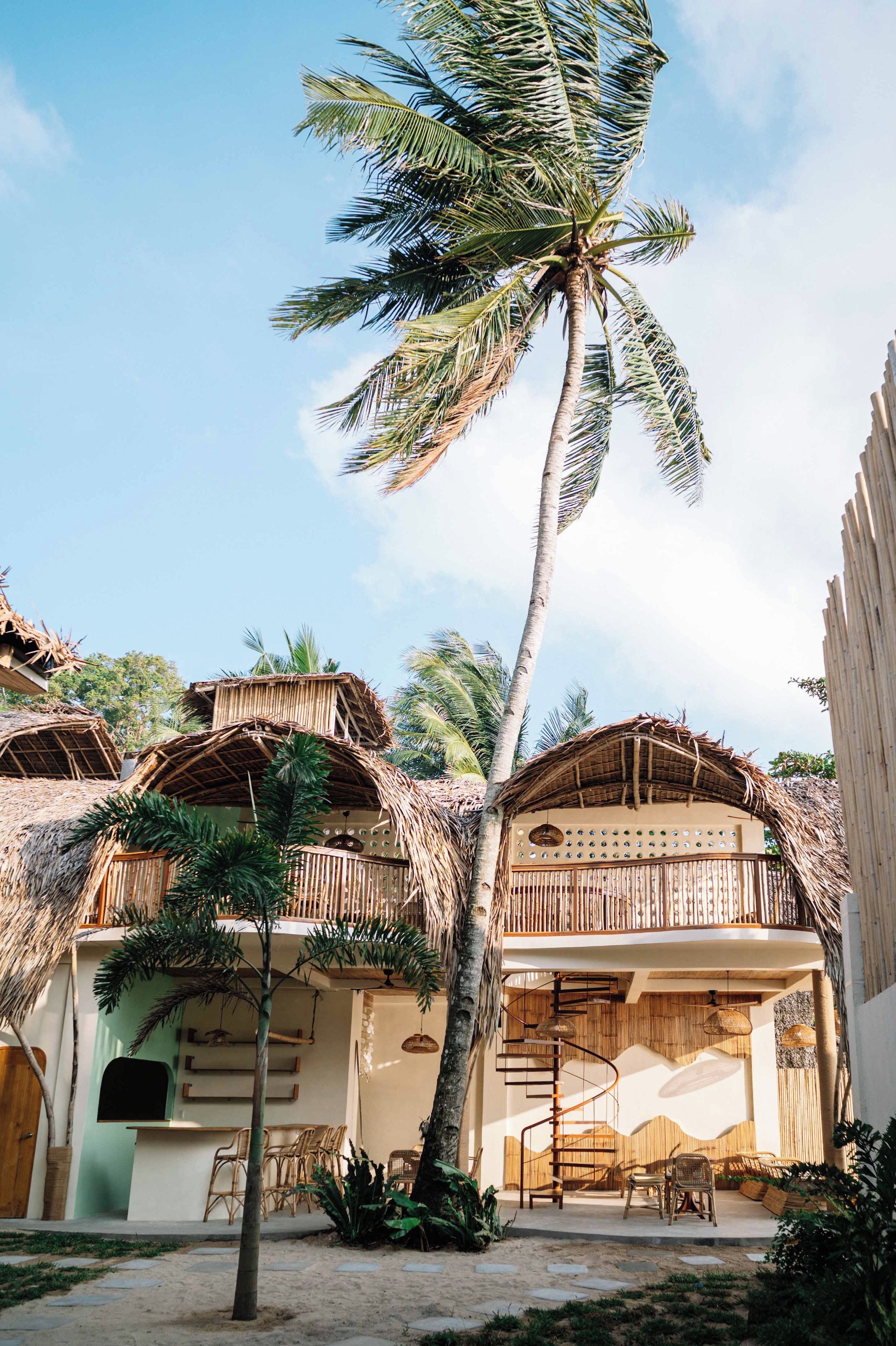 Resort tropical com construções de telhado de palha, uma palmeira e uma escada em espiral que leva a uma varanda.