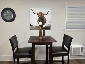 A dining area with a table and two chairs, a clock, a bull-themed artwork, and a window with blinds. - Peaceful Private Pet Friendly Studio-10 Miles from Buc-ee's & I65 (Harvest)