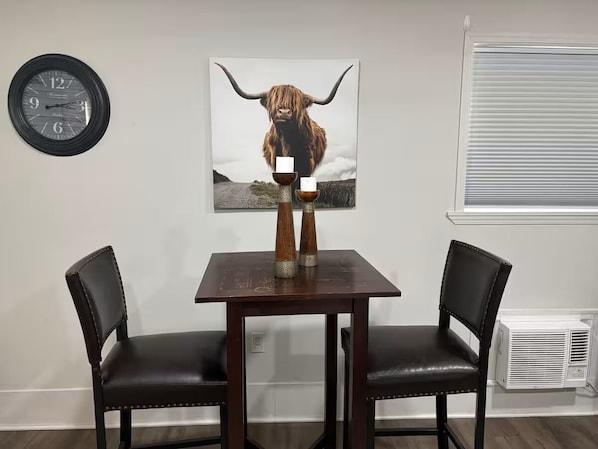A dining area with a table and two chairs, a clock, a bull-themed artwork, and a window with blinds.