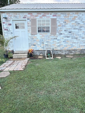A small house with a front door, a window with shutters, a garden with a potted plant, and a hose reel.