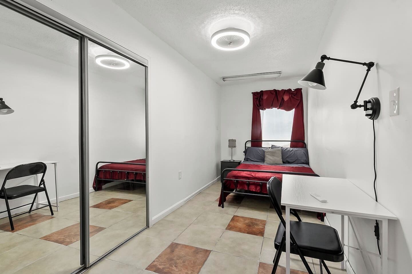 Een slaapkamer met een bed, een bureau, een stoel, een lamp en een spiegel.