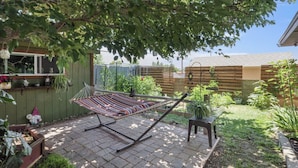 A hammock under a tree, a small table, and a garden with plants and a fence.