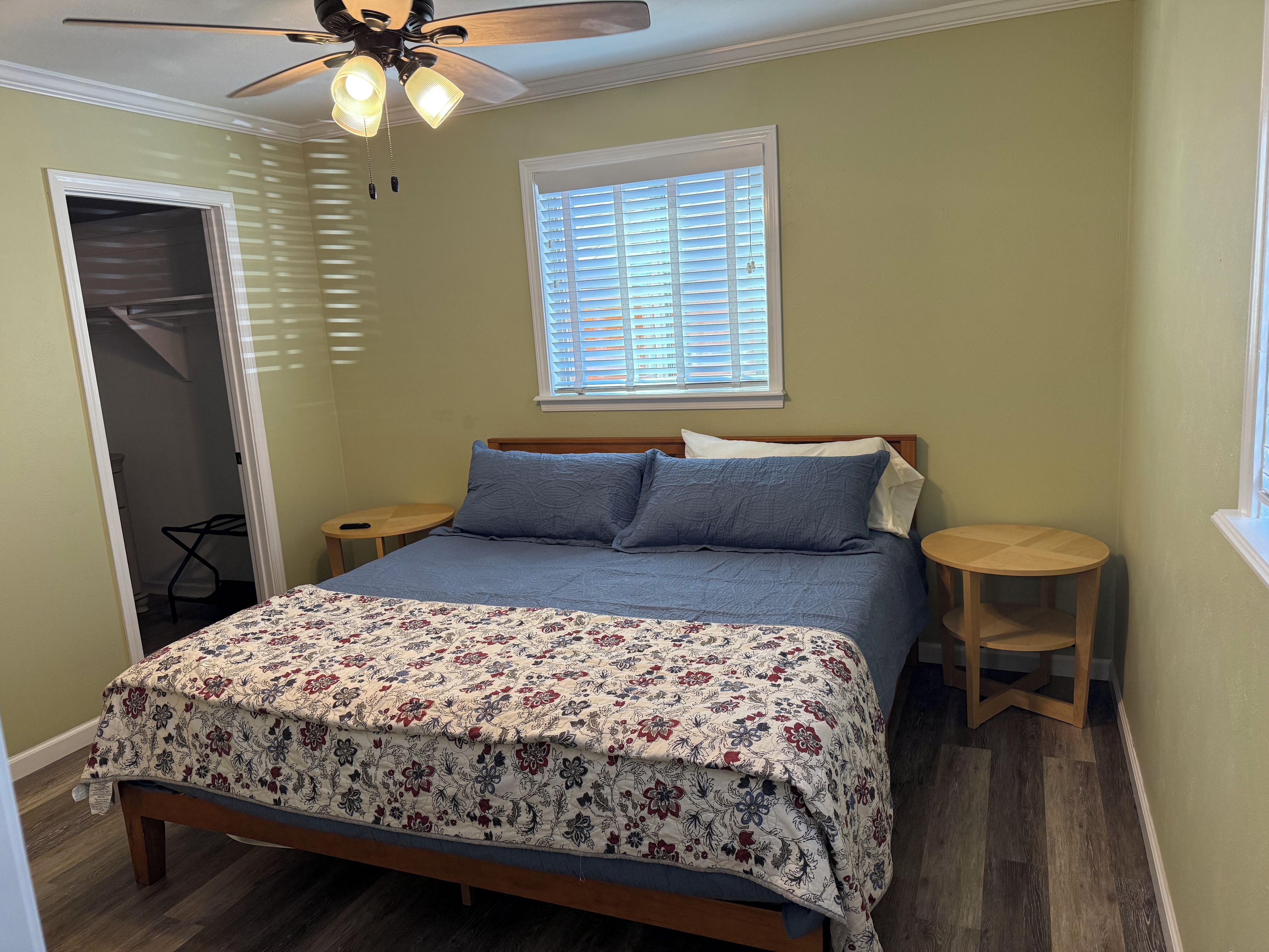 A bedroom with a bed, a ceiling fan, a window with blinds, a small table, and a chair.