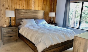Een slaapkamer met houten lambrisering, een bed, twee nachtkastjes en een raam met uitzicht op bomen.