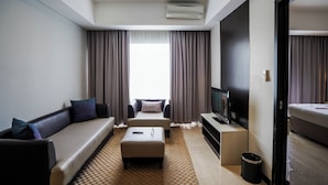 Habitación de hotel moderna con sofá, reposapiés, televisor de pantalla plana y un ventanal con cortinas.