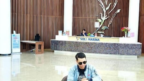 Un hombre sentado en una silla con una computadora portátil en un área de recepción con un mostrador identificado como 'SOLL MARINA'.