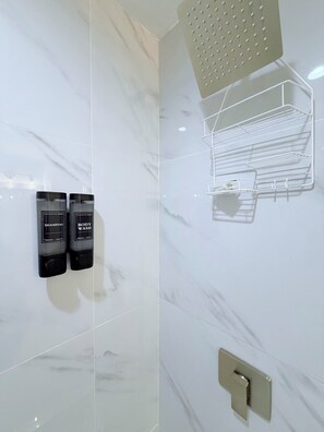 Une salle de bain avec un mur en marbre, une douche dotée d’un pommeau de douche à effet pluie, et deux bouteilles de shampoing et de gel douche.