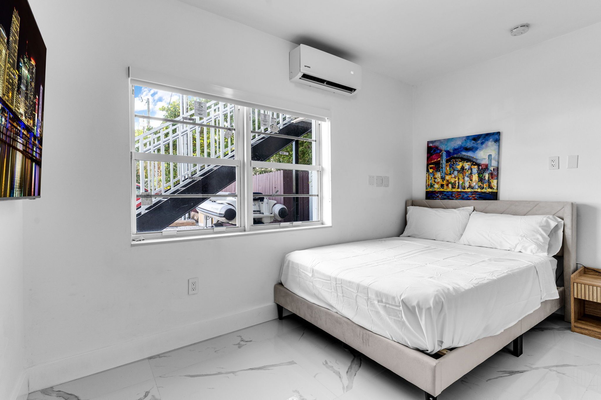 Une chambre à coucher avec un lit, une fenêtre donnant sur des immeubles, un appareil de climatisation et un tableau accroché au mur.