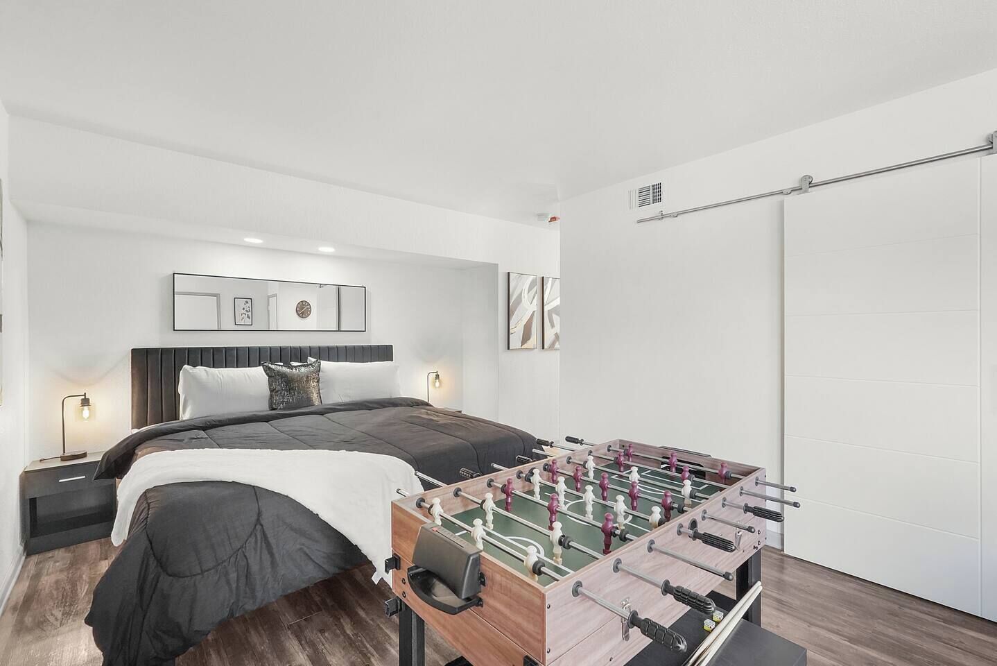 A modern bedroom with a bed, nightstands, a wall-mounted mirror, artwork, and a foosball table.