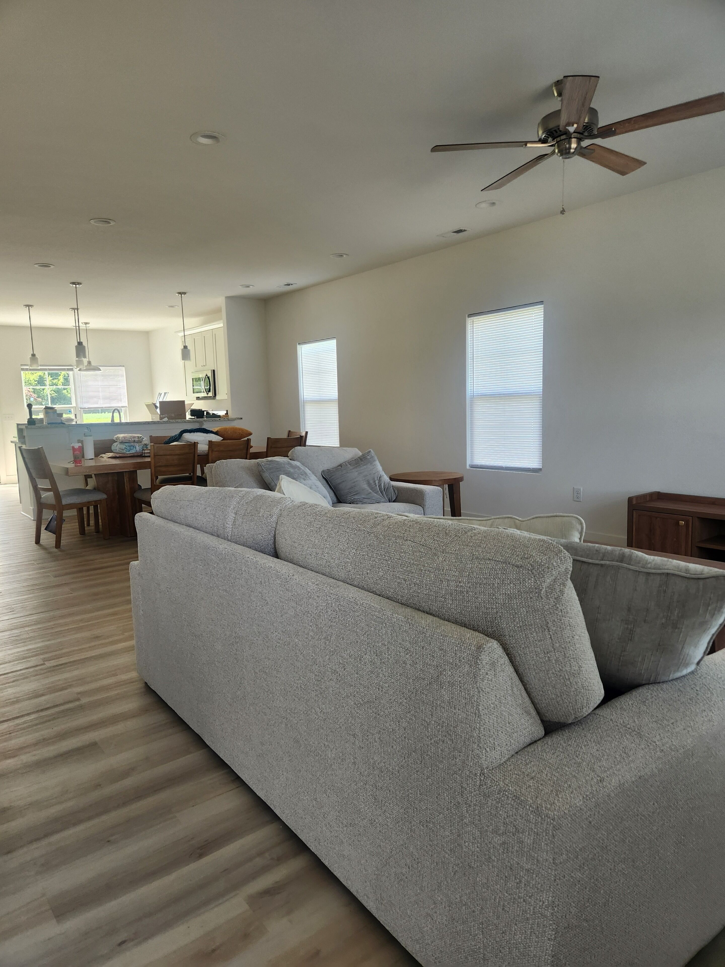 A spacious living area with a large gray sofa, dining table, and chairs. A ceiling fan is visible, and there are windows allowing natural light.