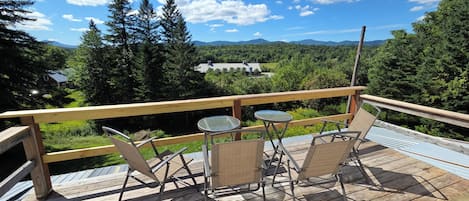 A wooden deck with outdoor furniture, including two chairs and a table, overlooking a forested landscape with mountains in the distance.