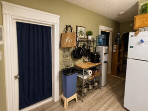 Una cocina con refrigerador, dispensador de agua, cafetera y estante con ollas y sartenes.