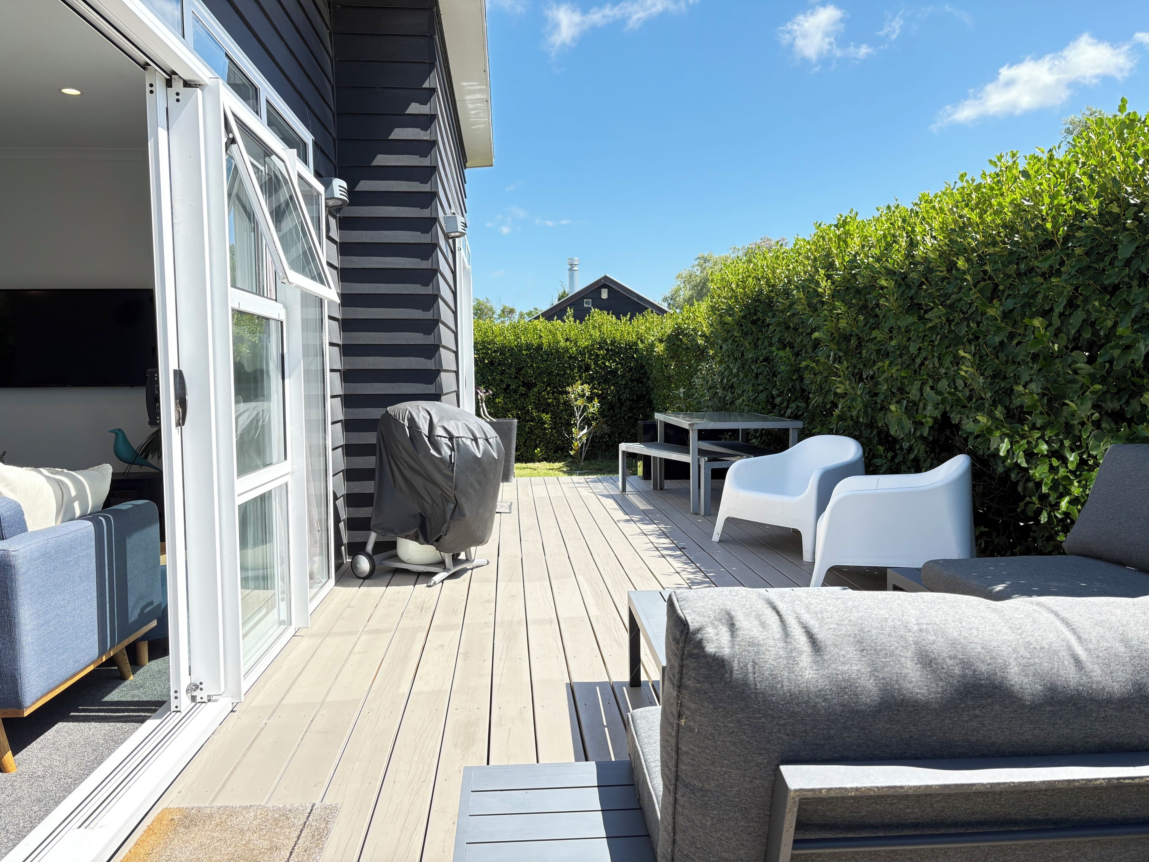 Een terras met buitenmeubilair, een overdekte barbecue en een uitzicht op een haag en een huis op de achtergrond.