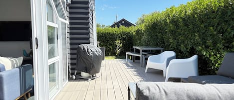 Una terraza con mobiliario exterior, una parrilla cubierta y vista a un seto y una casa al fondo.