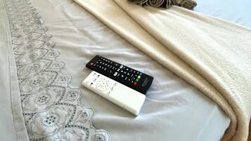 Cama bem arrumada com manta branca, toalha enrolada e controle remoto sobre a cama.