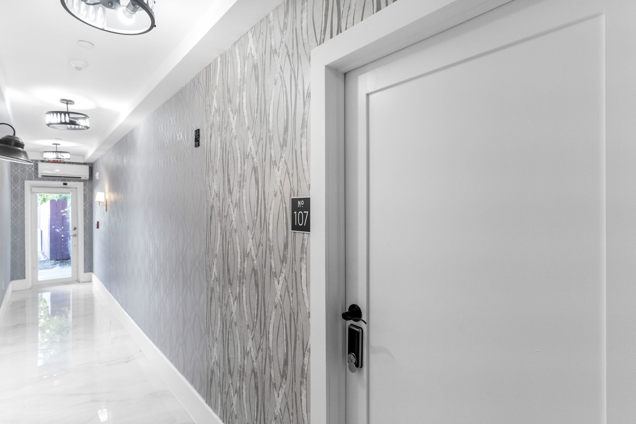 Un pasillo moderno con pared de diseño de mármol, una puerta blanca con placa de número y un piso elegante y reflectante.