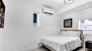 Un dormitorio con cama, unidad de aire acondicionado, ventana y mesita de noche.