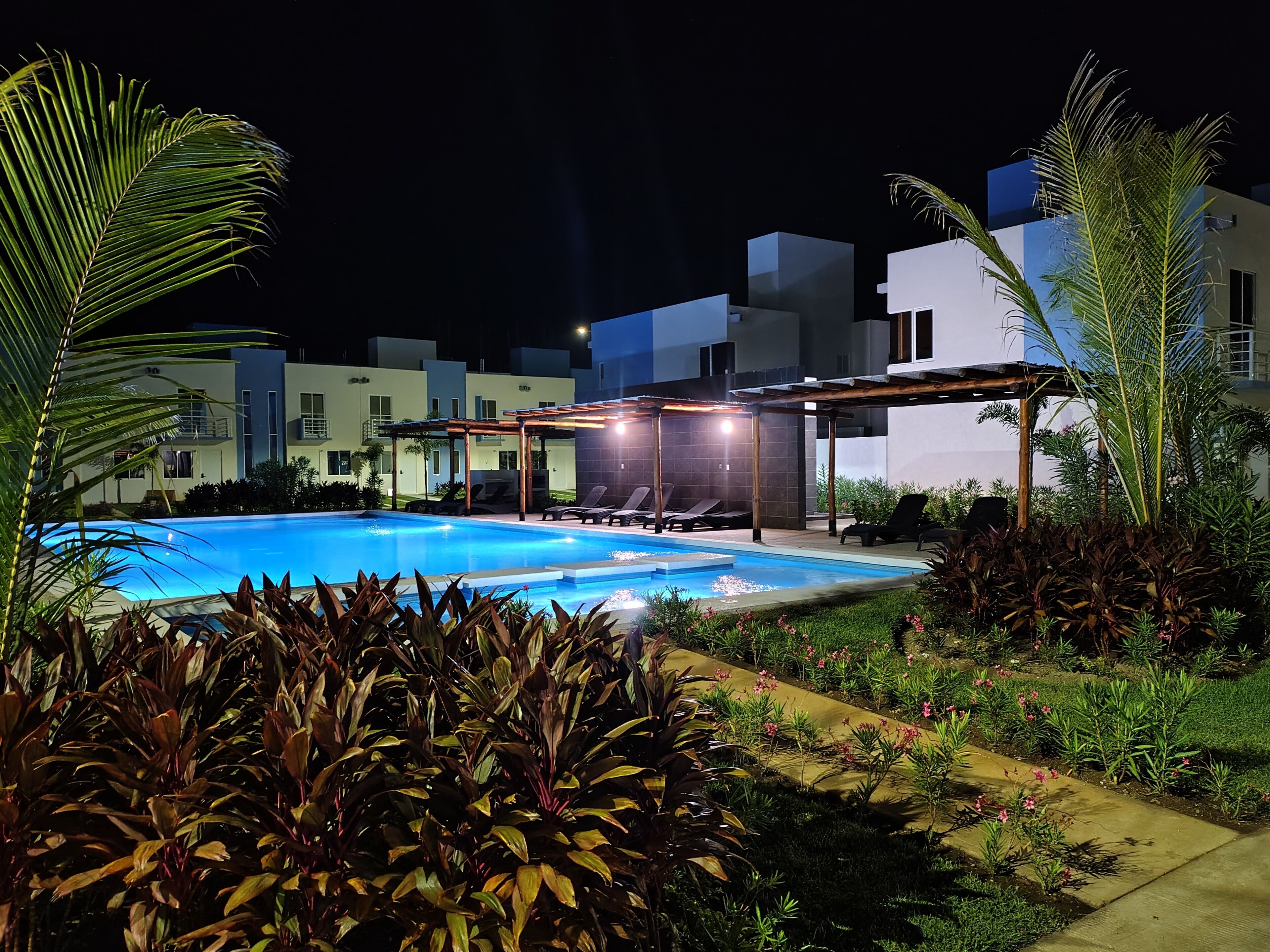 Une piscine entourée de chaises longues, de verdure et de bâtiments modernes, le tout illuminé la nuit.