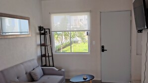 Een woonkamer met een grijze bank, een kleine tafel met een bezem, een televisie aan de muur en een deur.