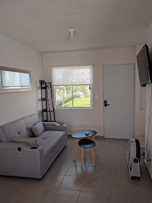 A living room with a grey sofa, a small table with a broom, a TV on the wall, and a door. - CASA MARIEL ACAPULCO DIAMANTE 🌊🌴 POOL, GARDEN, BEACH, SUN, SAND, AND OCEAN (Acapulco de Juárez)