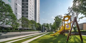 Un terrain de jeux avec un toboggan jaune et des balançoires, entouré d’une pelouse bien entretenue et d’arbres, avec un grand immeuble en arrière-plan.