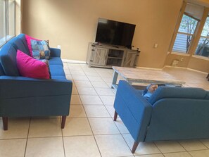 A living room with a blue sofa, a television on a wooden stand, and a window with blinds. - 4BR Denton Home Near UNT & TWU--Perfect for Work Trips & Families (Texarkana)