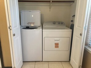 A laundry room with a front-loading washer and dryer, a small shelf above the washer, and a door with a metal handle. - 4BR Denton Home Near UNT & TWU--Perfect for Work Trips & Families (Texarkana)