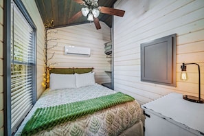 A bedroom with a bed, a ceiling fan, an air conditioning unit, a bedside lamp, and a window with blinds.