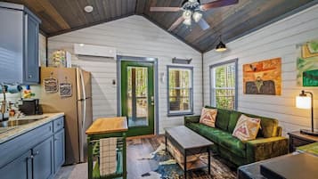 A compact kitchen with a refrigerator, sink, and wooden cabinets. There is a green door leading to a patio, and a green sofa with cushions in the living area.