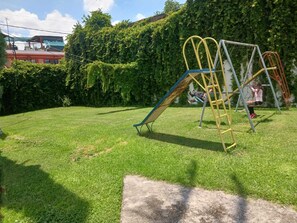 A playground with a slide and swings, surrounded by a grassy area and dense foliage.