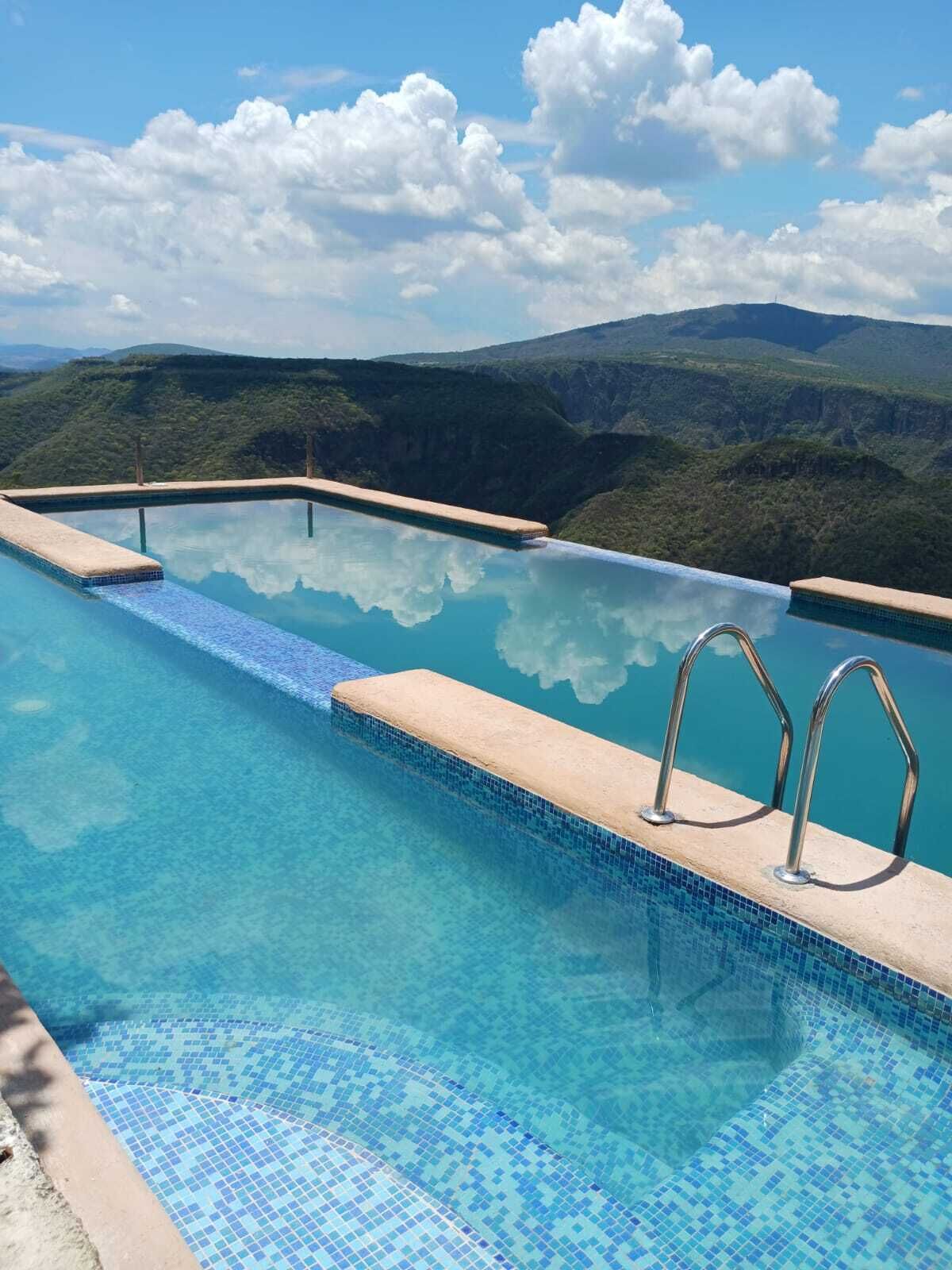Une magnifique piscine à débordement d’un bleu profond, bordée de carreaux, entourée d’un paysage naturel composé de collines et d’un ciel dégagé.