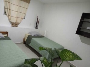 A small, simple room with two single beds, a window with a curtain, and a plant in the foreground.